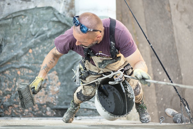 A man splattered with paint and plaster and held aloft by a rope harness performs restoration and repairs to the exterior of a commercial building.