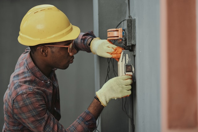 A male electrician with a yellow hard hat, safety glasses, and safety gloves works on an office's external electrical connection. Image at PrimeOfficeSpace.co.uk.