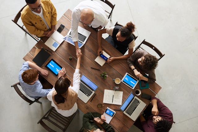 Top-down view of a group of colleagues sitting around a meeting room table. A woman at the centre of the table is standing and shaking hands with a man across the table from her. Image at PrimeOfficeSpace.co.uk.