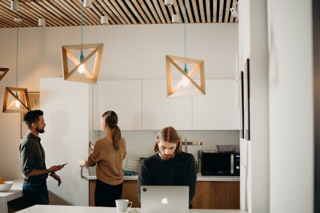 A man sits at a flexible workspace's kitchen island working on a silver laptop with a mug of coffee. A man and a woman are having a discussion behind him while she opens a cupboard. Image at PrimeOfficeSpace.co.uk.