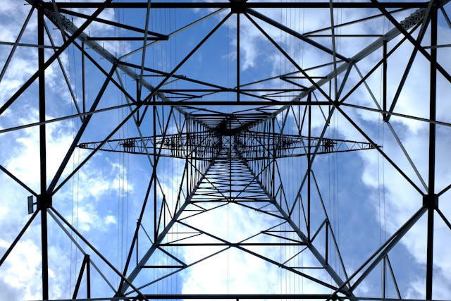 A bottom-up view from the base of a power line tower during the daytime. Behind the tower's intricate framework is a blue, cloud-strewn sky. Image at PrimeOfficeSpace.co.uk.