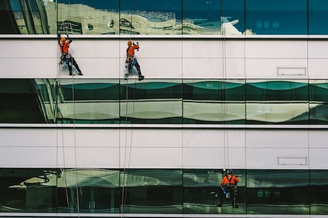 Three men in orange high-visibility shirts and hard hats clean external windows of an office building while being suspended by ropes and harnesses. Image at PrimeOfficeSpace.co.uk.