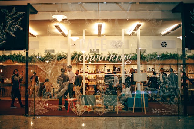 A view through the glass front of a busy coworking café with many people standing around and talking or sitting and working. Image at PrimeOfficeSpace.co.uk.