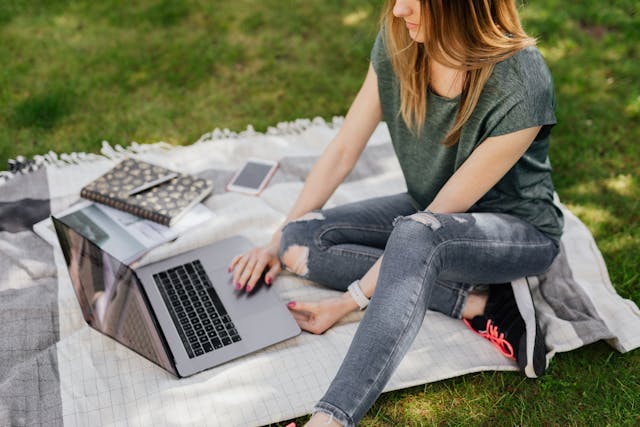 A woman wearing jeans, trainers, and a green t-shirt sitting on a picnic blanket, working on her laptop in a park. Image at PrimeOfficeSpace.co.uk.