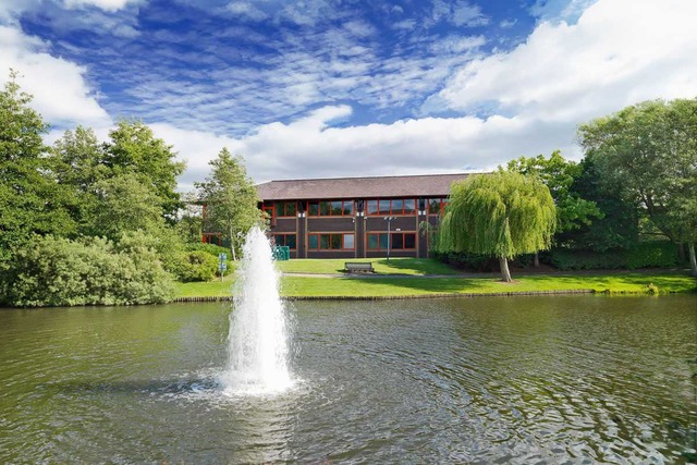 A midday view across a large pond towards a two-storey reddish office building at Aztec West Business Park in Bristol. A fountain spurts water from the centre of the pond, and many large trees surround the building on the lawn in front of the building. Image at PrimeOfficeSpace.co.uk.