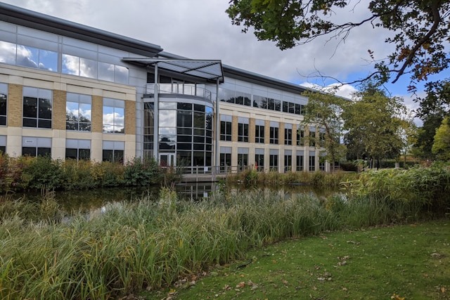A view of one of Birmingham Business Park’s many office buildings. There is a long pond in front of the building that seems to run its length. The building is three storeys tall, and its many windows reflect a cloudy sky and the many trees and long grass around the pond that also feature nearby. Image at PrimeOfficeSpace.co.uk.