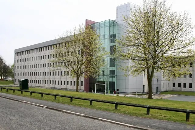 A view from the road towards a grey concrete office building at Birchwood Park business park in Warrington. The building is five storeys tall and has a greenish glass fronted foyer. Two trees with sparse leaves flank the entrance on the lawn around the building. Image at PrimeOfficeSpace.co.uk.