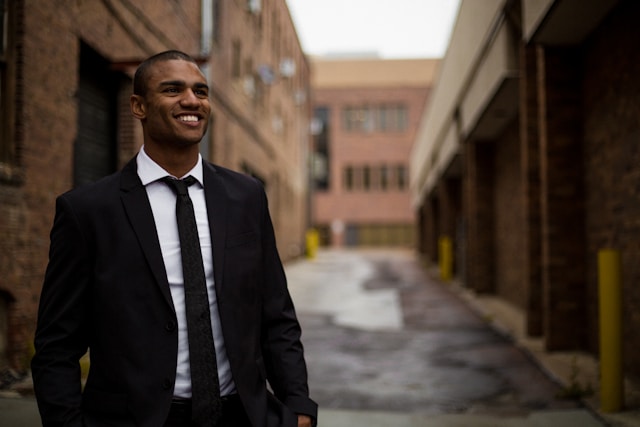 A businessman in a smart black suit with a bright white collared shirt and black tie stands in an alley between two red brick office buildings and smiles broadly. Image at PrimeOfficeSpace.co.uk.