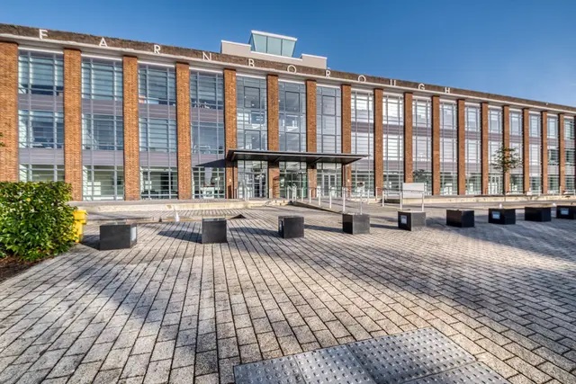 A view from the wide, brick-paved path leading to the front of the Farnborough Business Park’s main building, which has the word “Farnborough” spelled out in wide-spaced letters as signage across the front of the roof of the three-storey building. Image at PrimeOfficeSpace.co.uk.