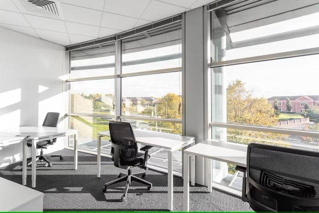 A daytime view from within an upper level office at Chester Business Park in Cheshire, outside the floor-to-ceiling windows towards the rest of the business park. A couple of white desks with black office chairs sit against the windows. Image at PrimeOfficeSpace.co.uk.