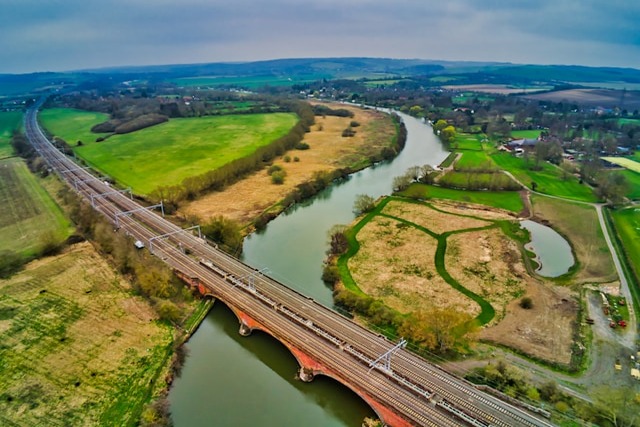 A daytime aerial view over the River Thames winding its way through Oxfordshire. A train line is going across the river in the foreground and bends away to disappear in the distance too across green fields. Image at PrimeOfficeSpace.co.uk.