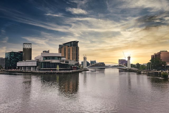 A dusk view over the water at Salford Quays, which borders Trafford Park business park. Building line the waterway and a white suspension bridge spans the two banks. The sun is near the horizon in the distance behind the bridge, dramatically lighting the sky filled with wispy clouds. Image at PrimeOfficeSpace.co.uk.