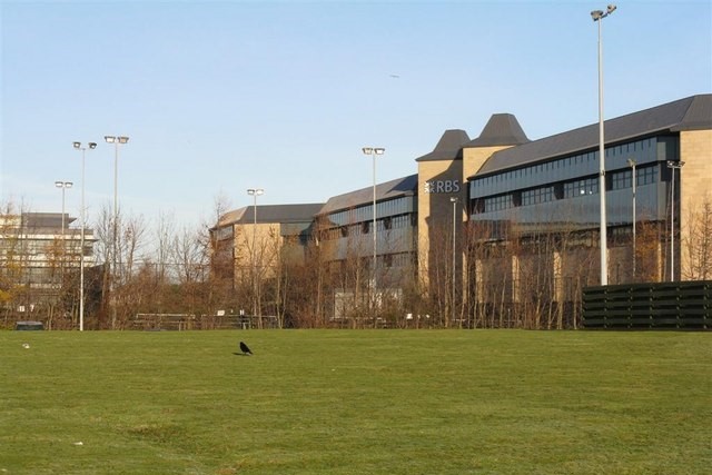 A daytime view from a grassy field towards one of the office buildings at South Gyle Trade Park business park in Edinburgh, Scotland. Many bare trees line the field between the field and the building. A lone black crow stands in the middle of the field. Image at PrimeOfficeSpace.co.uk.