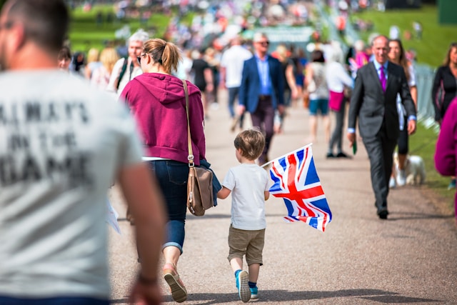 A sunny daytime view down a wide walkway with many people walking to and fro. A woman in a pink sweatshirt is in focus in the centre of the image, and she’s holding the hand of her young son, who’s holding a Union Jack flag. Image at LondonOfficeSpace.com.