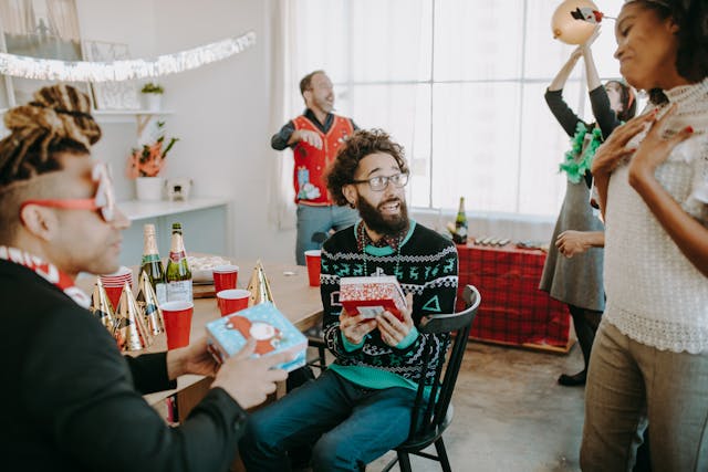 Five colleagues in colourful Christmas jumpers or smart casual attire are pictured at a festive season celebration. Two seated men are holding out gifts, and their colleague, a woman, is standing near them and looking very grateful for the gifts they have offered. Their two colleagues, a man and a woman, are playing with a balloon in the background, passing it to each other through the air. Image at PrimeOfficeSpace.co.uk.