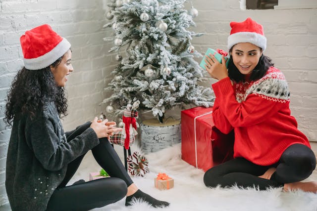 Two women colleagues wearing Christmas jumpers and red and white Santa hats are sitting on a fluffy white rug next to a white-dusted Christmas tree with silver baubles, exchanging gifts and smiling. Image at PrimeOfficeSpace.co.uk.