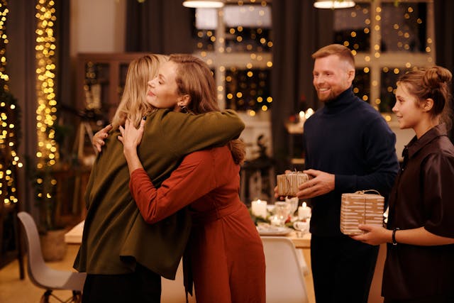 Two women are hugging at a tastefully decorated Christmas party, and their colleagues, a man and a woman, are smiling at them and holding wrapped gifts. Image at PrimeOfficeSpace.co.uk.