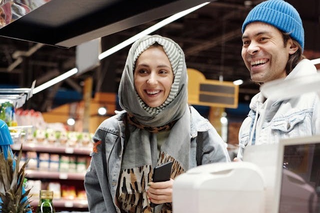 Two customers, a young woman and a young man, are smiling broadly in conversation with a shop worker who is processing their transaction at the checkout. The woman is wearing a grey hijab and grey denim jacket, and the man is wearing a bright blue beanie and light denim jacket over a white hoodie. A few of their items are still on the checkout conveyor, yet to be scanned, like a pineapple and bottle of cooking oil, and full shelves of dry goods are visible in the background. Image at PrimeOfficeSpace.co.uk.