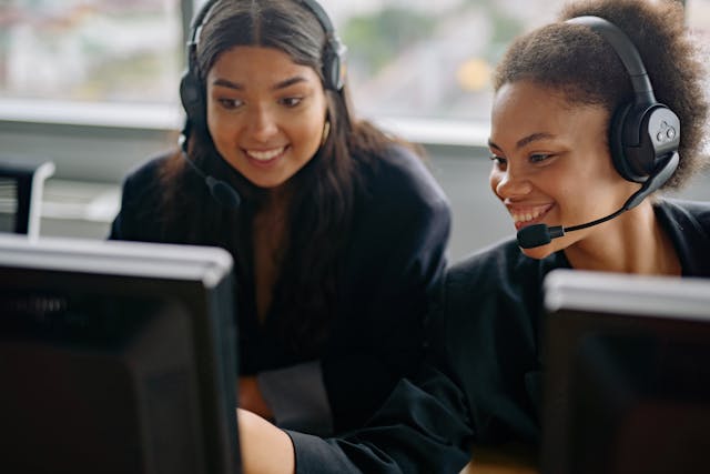 Two women customer service agents are sitting next to each other at their desks and smiling. They are both wearing black headsets with the microphones extending out in front of their mouths, and the woman on the right is leaning across to point at something on her colleague’s computer monitor. Image at PrimeOfficeSpace.co.uk.