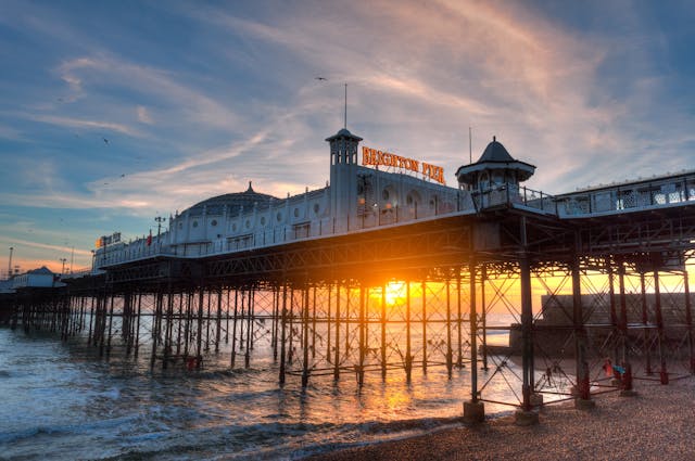 A view from the shore up at the famous Brighton Pier at sunset with the bright orange setting sun peeking between the pier's support beams. Image at PrimeOfficeSpace.co.uk.