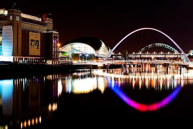 A view along Newcastle's River Tyne at night with many lights reflecting on the black river surface. The historic Baltic Flour Mills building now housing the Baltic Centre for Contemporary Art and the shiny, slug-like building, The Sage, are on the left bank and the iconic Gateshead Millennium Bridge is lit up in neon and spans the river. Image at PrimeOfficeSpace.co.uk.