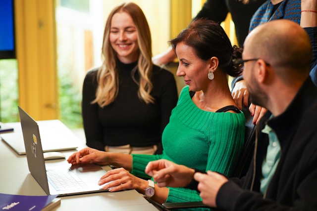 A woman in a bright green knitwear top is sitting at a desk with her laptop in front of her. Her startup colleagues, a man and a woman wearing black tops, sit on either side of her smiling, and another of their male colleagues stands behind them, resting his hands on the backs of their chairs. Image at PrimeOfficeSpace.co.uk.