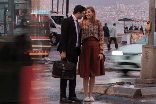 A man in a black business suit and carrying a briefcase holds hands with his female colleague in a brown and tan patterned cardigan and a brown skirt. They are standing next to a busy city street at dusk after rain with cars and people milling about and much of the large city can be seen spread out in the background across hills. Image at PrimeOfficeSpace.co.uk.