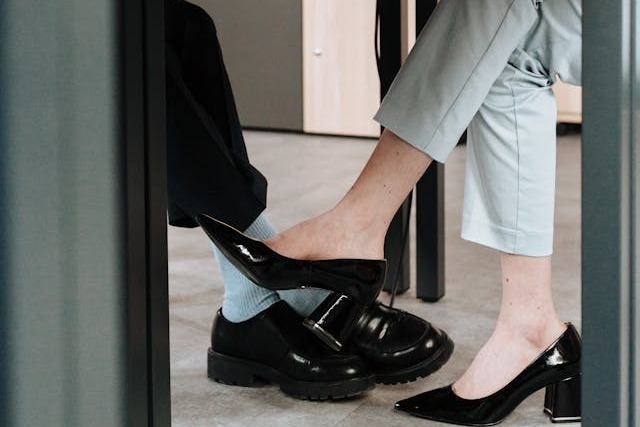 Close-up view under an office desk where two coworkers, a man and a woman, sit across from each other playing footsie. The woman, who is wearing light grey business pants and black high heels, is rubbing one of her feet on the man’s lower leg. He is wearing shiny, black leather shoes, light blue socks, and black business pants. Image at PrimeOfficeSpace.co.uk.