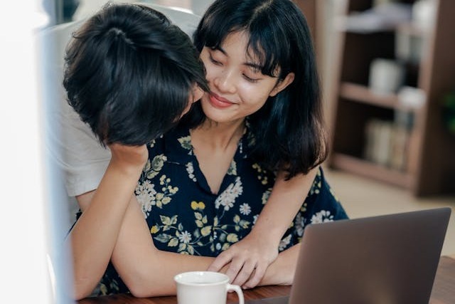 A businessperson with short black hair and wearing a short-sleeved white collared shirt is standing behind their female colleague and putting their arms around her as she sits at her desk with her laptop and mug of coffee on the desk in front of her. She is cradling her colleague’s head in her hand as they lean over her, and she is smiling affectionately. Image at PrimeOfficeSpace.co.uk.