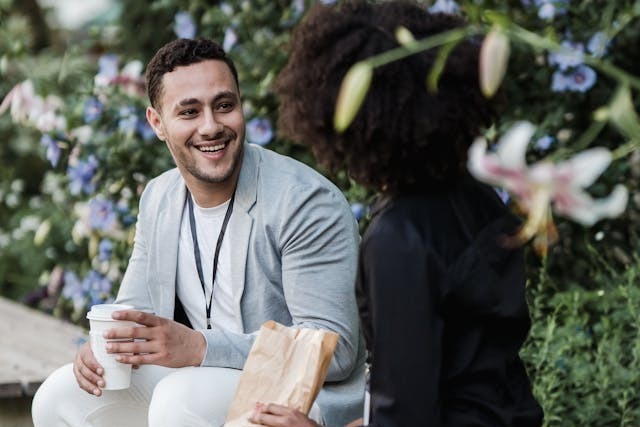 Two coworkers, a man and a woman, are sitting next to each other on a bench outside on their lunch break. The man wears a light grey blazer with a white t-shirt and pants with his work ID hanging from a lanyard around his neck. He is holding a coffee cup and smiling while looking at the woman beside him, who is dressed in black and holding a small brown paper bag from a bakery. Flowers of different colours and lush green bushes surround them. Image at PrimeOfficeSpace.co.uk.